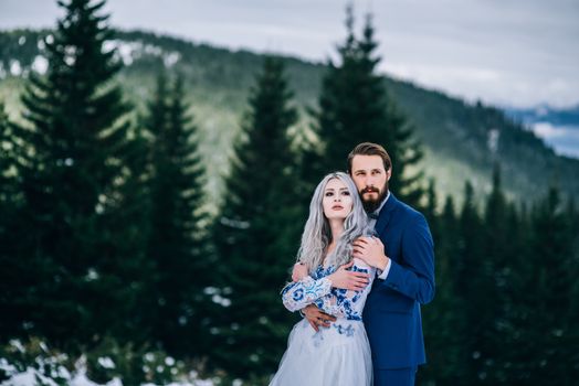 groom in a blue suit and bride in white, embroidered with blue pattern, dress on a background of green pine forests in the mountains of the Carpathians