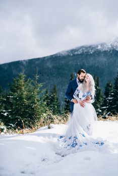 groom in a blue suit and bride in white, embroidered with blue pattern, dress on a background of green pine forests in the mountains of the Carpathians