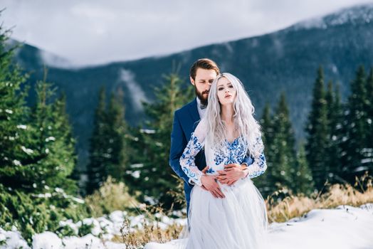 groom in a blue suit and bride in white, embroidered with blue pattern, dress on a background of green pine forests in the mountains of the Carpathians