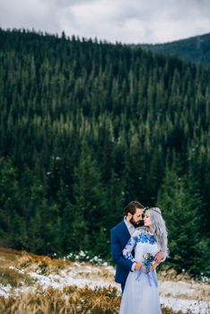 groom in a blue suit and bride in white, embroidered with blue pattern, dress on a background of green pine forests in the mountains of the Carpathians