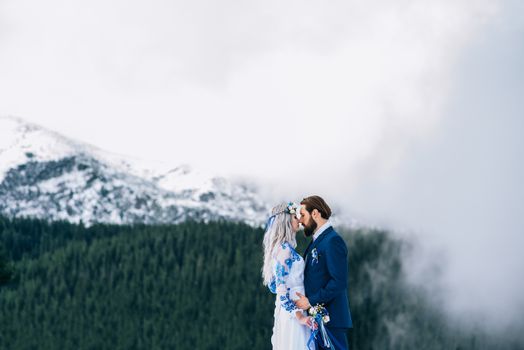 groom in a blue suit and bride in white, embroidered with blue pattern, dress on a background of green pine forests in the mountains of the Carpathians