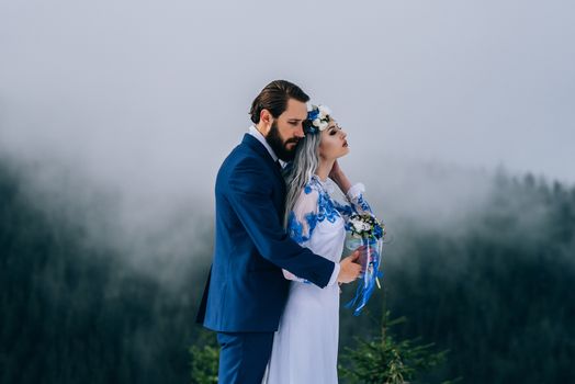 groom in a blue suit and bride in white, embroidered with blue pattern, dress on a background of green pine forests in the mountains of the Carpathians