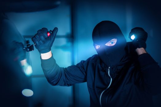 Car Thief Car Robbery Concept Photo. Caucasian Male Thief in Black Mask Trying to Open Car Using Custom Tool and Flashlight. Car Robber.