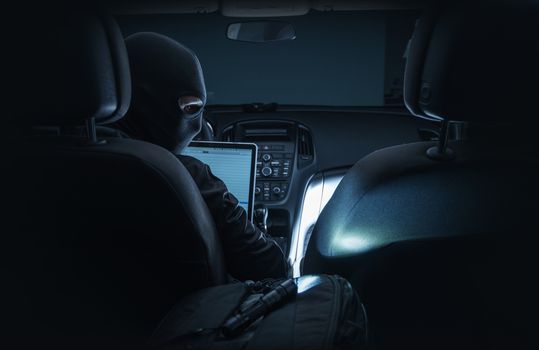 Hacking Car System. Car Hacker in Black Mask Hacking Vehicle Systems From Inside the Car Using Laptop Computer.