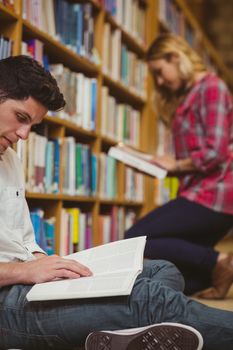 Smiling classmates reading book while leaning on bookshelves in library