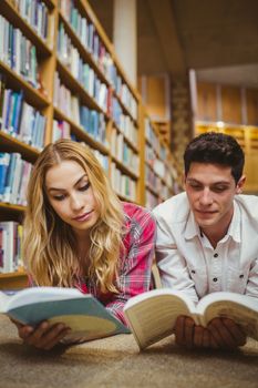 Smiling classmates reading book while leaning on bookshelves in library