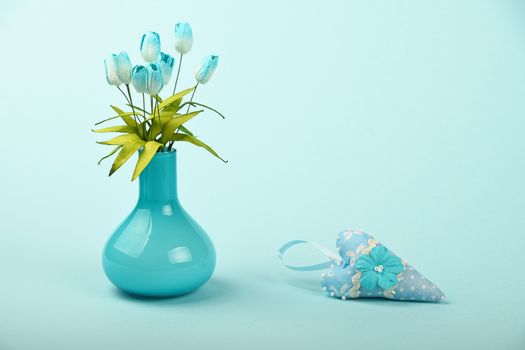One small textile toy heart and mulberry silk tulip flowers bouquet in glass vase on tender blue design paper background
