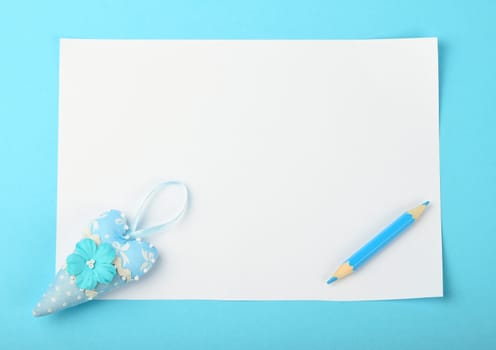 White paper note with tiny textile toy heart and small pencil on tender blue background