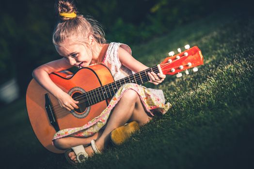 Little Girl Playing Acoustic Guitar While Seating on the Backyard Grass. 