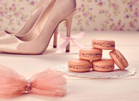 Woman essentials fashion accessories. Macarons french dessert, luxury beige shoes high heels, bow. Creative glamor wedding set, vanilla wood, floral background. Romantic, still life. Retro vintage 