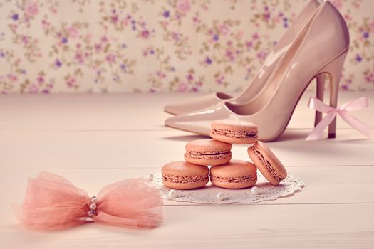 Woman essentials fashion accessories. Macarons french dessert, luxury beige shoes high heels, pearl, bow. Creative wedding set, vanilla wood, floral background. Romantic, still life. Retro vintage 