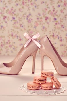 Woman essentials fashion accessories. Macarons french dessert, luxury beige shoes high heels, pearl, bow. Creative wedding set, vanilla wood, floral background. Romantic, still life. Retro vintage 
