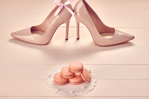 Woman essentials fashion accessories. Macarons french dessert, luxury beige shoes high heels, pearl, bow. Creative glamor wedding set, vanilla wood background. Romantic, still life. Retro vintage 