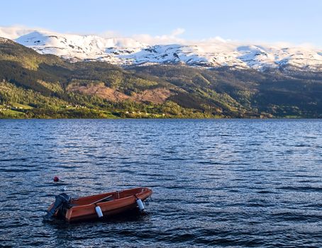 Red motorboat on a lake. On the other side houses and snowy mountain range in the rays of the evening sun. Water is blue, wavy. Seagull sitting on the boat. Voss, Norway