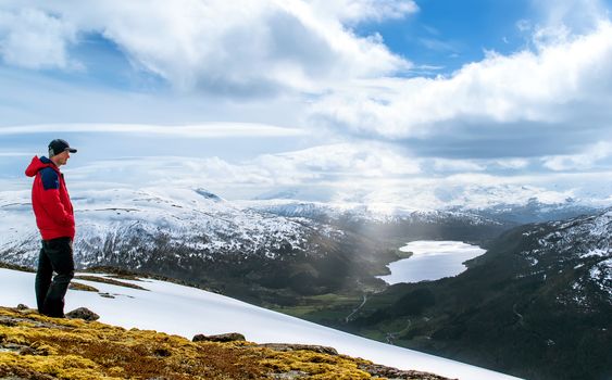 Tourist stands on a top and looks into the distance. Lake in the valley.  Blue sky with clouds. Snowy mountains. Sun rays breaks through the clouds. Norway.