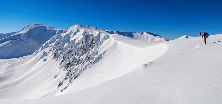 Panorama of snowy mountain peaks. Sharp rocks on a steep slope. Two tourists. The sky is clear, sunny. Winter. Ukraine