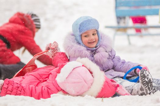 Two girls in the winter ride on a snowy hill surrounded by other children