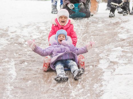 Two girls in the winter ride on a snowy hill surrounded by other children