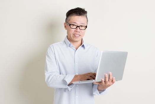 Portrait of single mature 50s Asian man in casual business using laptop pc and smiling, standing over plain background with shadow. Chinese senior male people.