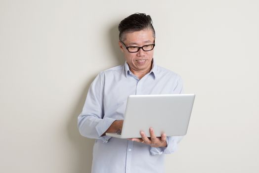 Portrait of modern mature 50s Asian man in casual business using notebook computer and smiling, standing over plain background with shadow. Chinese senior male people.