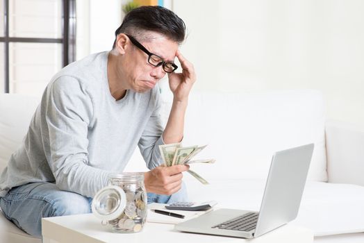 Financial problem concept. Portrait of 50s mature Asian man counting money with worried expression, sitting on sofa at home.