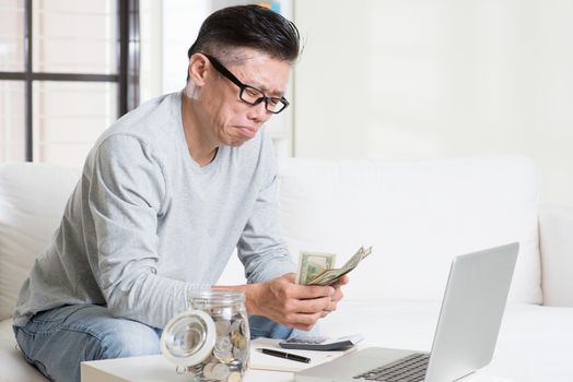 Financial problem concept. Portrait of 50s mature Asian man counting money with worried expression, sitting on sofa at home.