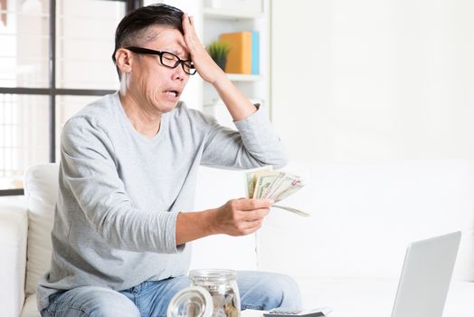 Financial crisis concept. Portrait of 50s mature Asian man counting money with sad expression, sitting on sofa at home.