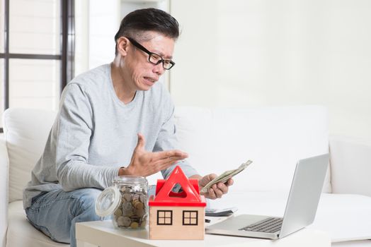 Cash flow problem concept. Portrait of 50s mature Asian man counting money with sad expression, sitting on sofa at home.