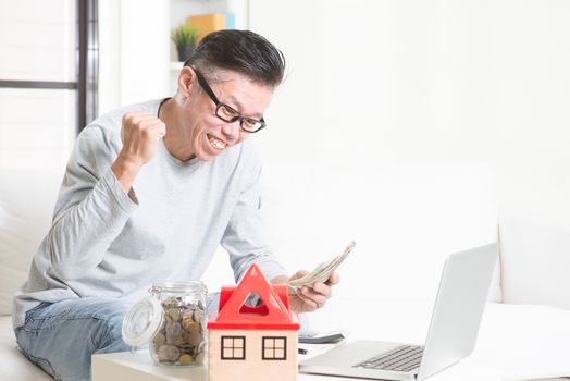 Portrait of happy 50s mature Asian man counting on money and smiling. Saving, retirement, retirees financial planning concept. Family living lifestyle at home.