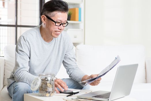 Portrait of 50s mature Asian man looking at bill and laptop in the living room. Saving, retirement, retirees financial planning concept.