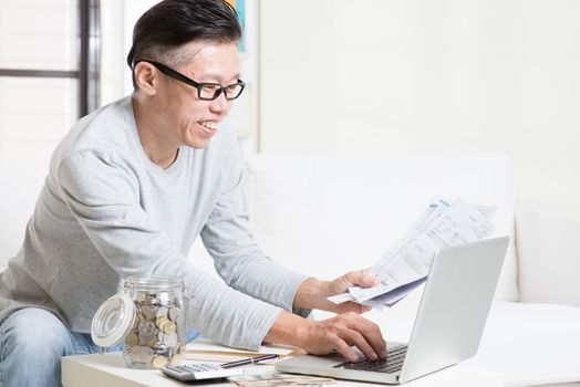 Portrait of 50s mature Asian man looking at laptop and paying bills online in the living room. Saving, retirement, retirees financial planning concept.