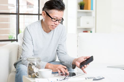 Mature 50s Asian man doing analysis on his budget, looking on tablet pc and smart phone. Saving, retirement, retirees financial planning concept.