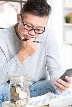 Mature 50s Asian man doing analysis on his financial investment, looking on data chart and smartphone. Saving, retirement, retirees financial planning concept.