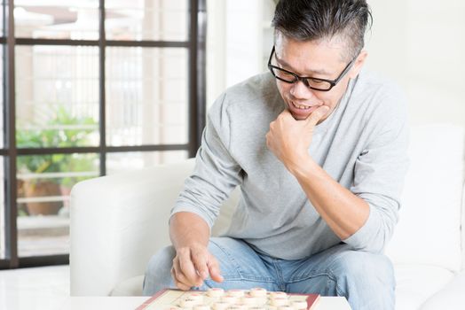 Mature 50s Asian man playing China chess, sitting on couch at home.