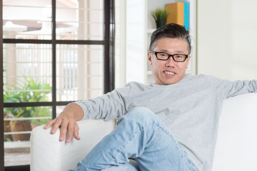 Portrait of mature 50s Asian man smiling and sitting at home. Senior Chinese male relaxed and seated on sofa indoor.