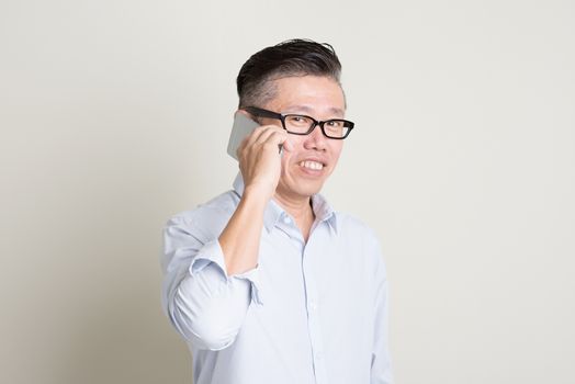 Portrait of single mature 50s Asian man in casual business making a call using smartphone and smiling, standing over plain background with shadow. Chinese senior male people.