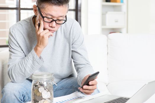 Mature 50s Asian man doing analysis on his spending, looking on data chart and smart phone. Saving, retirement, retirees financial planning concept.