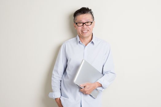 Portrait of modern mature 50s Asian man in casual business holding laptop computer and smiling, standing over plain background with shadow. Chinese senior male people.