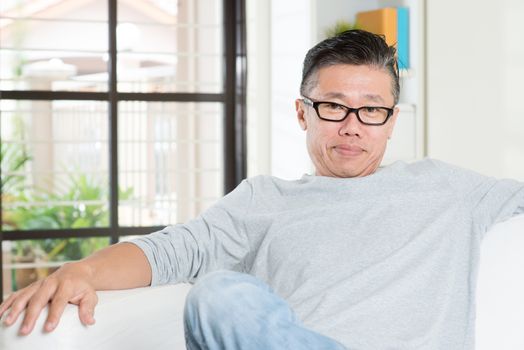 Portrait of mature 50s Asian man sitting at home. Senior Chinese male relaxed and seated on sofa indoor.