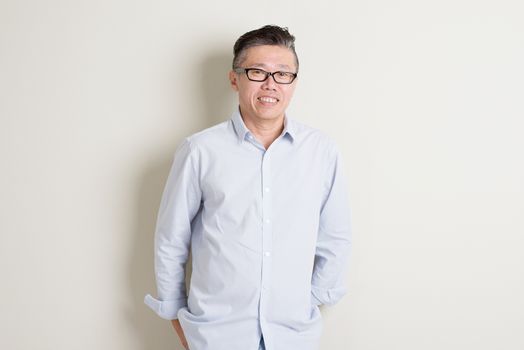 Portrait of single mature 50s Asian man in casual business arms crossed and smiling, standing over plain background with shadow.