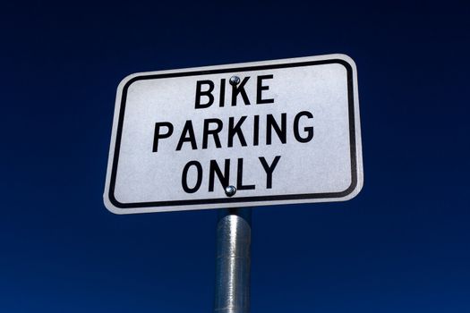 Bicycle parking only sign with black letters on white background and blue sky.