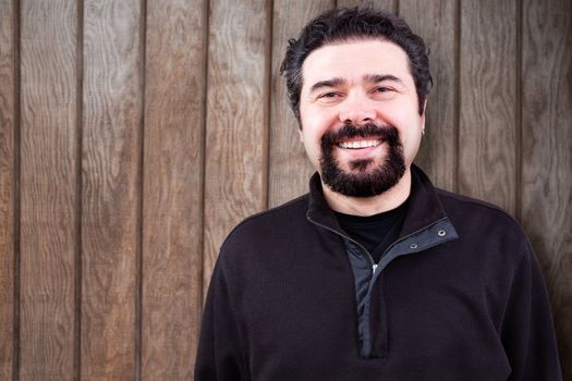 Half Body Shot of a Middle Aged Man with Goatee Beard, Sincerely Smiling at the Camera, Against Wooden Wall with Copy Space.