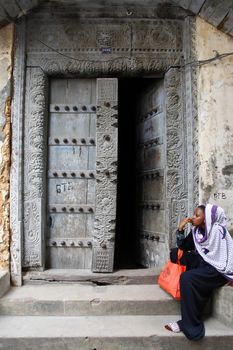 Stone Town, Tanzania - January 1, 2016: Traditional house with old door and woman in traditional Muslim clothes sitting on the street of Stone Town.