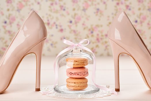 Woman essentials fashion accessories. Macarons french dessert, luxury beige shoes high heels, pearl, ribbon. Creative wedding set, vanilla wood, floral background. Romantic, still life. Retro vintage 