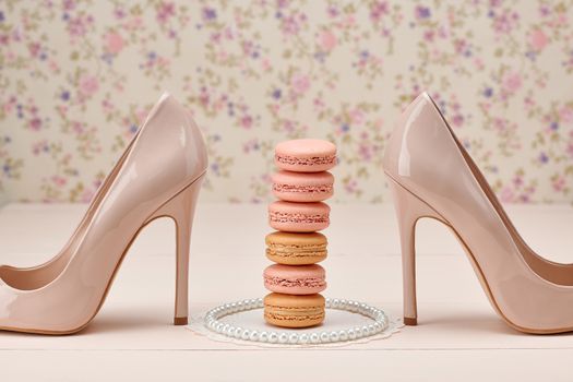 Woman essentials fashion accessories. Macarons french dessert, pearl necklace, luxury beige shoes high heels. Creative wedding set, vanilla wood, floral background. Romantic, still life.Retro vintage 