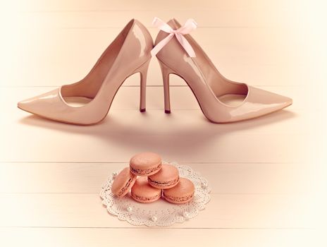 Woman essentials fashion accessories. Macarons french dessert, luxury beige shoes high heels, bow. Creative glamor wedding set, vanilla wood background. Romantic, still life. Retro vintage 