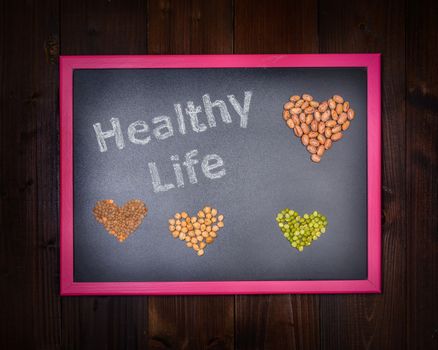 In the picture a blackboard that says "Healthy Life" and around a set of hearts made from chickpeas, beans, lentils and peas on wooden background