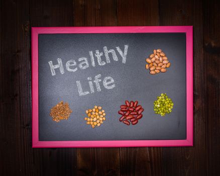 In the picture a blackboard that says "Healthy Life" and around small groups made up of chickpeas, beans, lentils and peas on wooden background