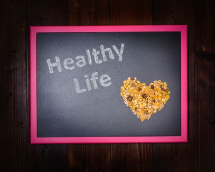 In the picture a blackboard, on the left side with written "Healthy Life" and on the right side a mixture of cereals which draw a heart on wooden background