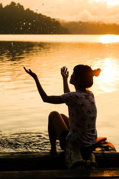 Silhouetted Thai girl sitting on a wood raft is splashing water in the air with background of dam and sunrise at Suratthani Province of Thailand